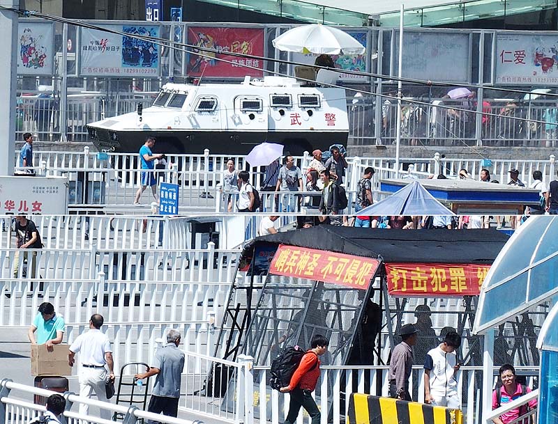 装甲車が陣取るウルムチ南駅前(上)と金網越しで銃剣装備のライフルを構える武警 800